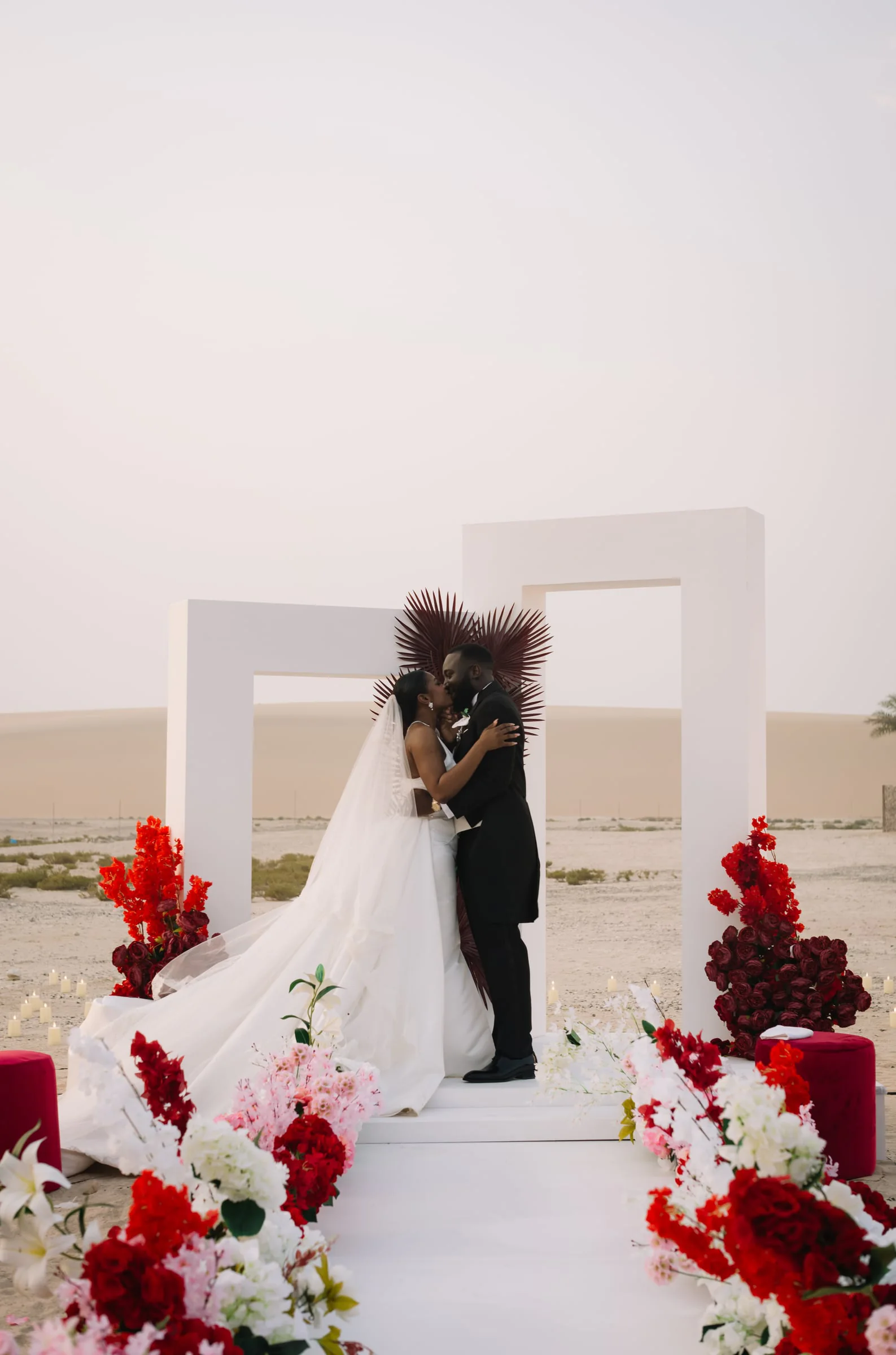 Bride and groom kissing beneath a geometric arch surrounded by red florals
